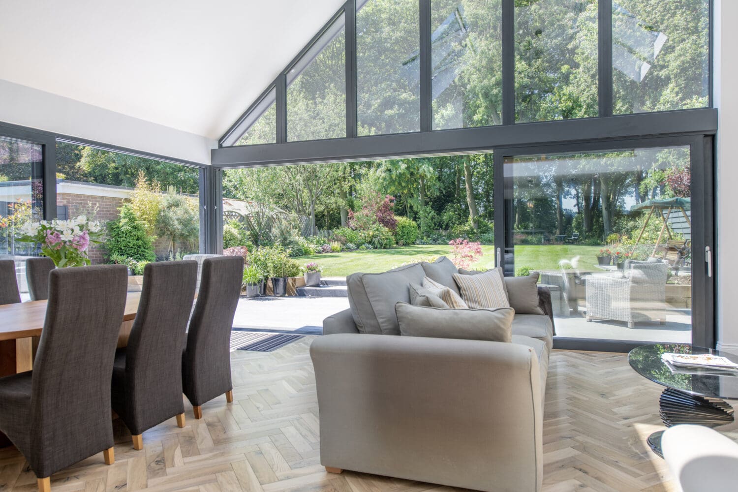 A modern living and dining area with large floor-to-ceiling windows and sliding doors opens to a lush green garden and patio with outdoor furniture. Natural light fills the space, highlighting wooden floors and neutral furniture.