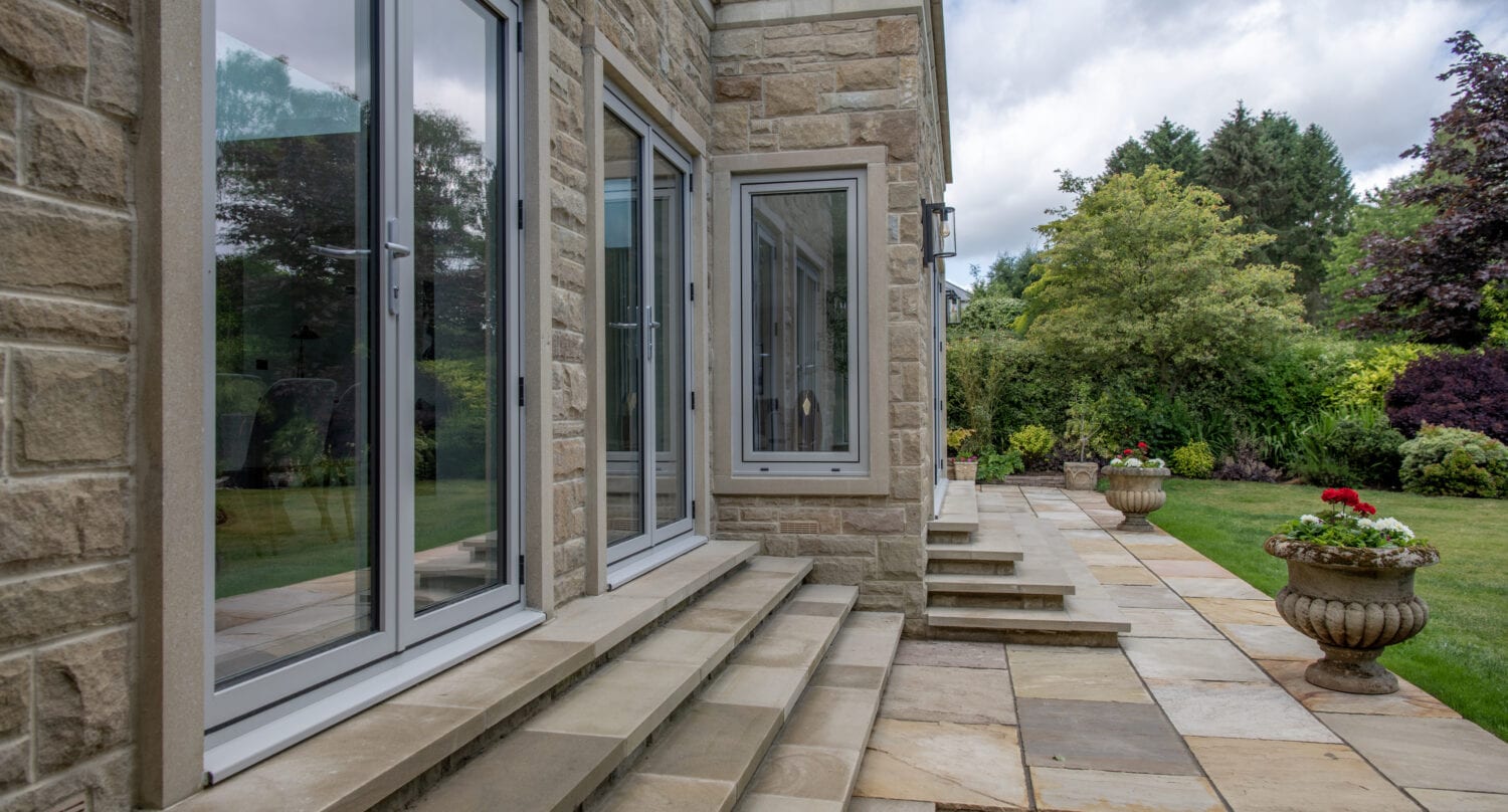 Stone patio steps lead to sliding doors on a modern stone house, overlooking a landscaped garden with large planters, green lawn, trees, and cloudy sky.