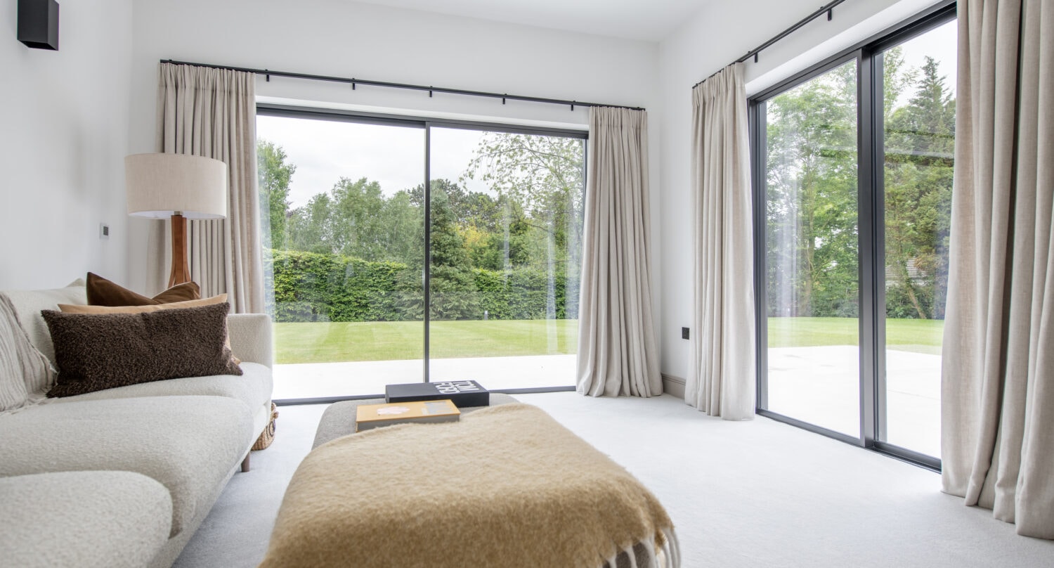 A bright modern living room with bifold doors, beige curtains, a white sofa with brown cushions, a floor lamp, and a tan throw blanket on an ottoman, overlooks a green garden.
