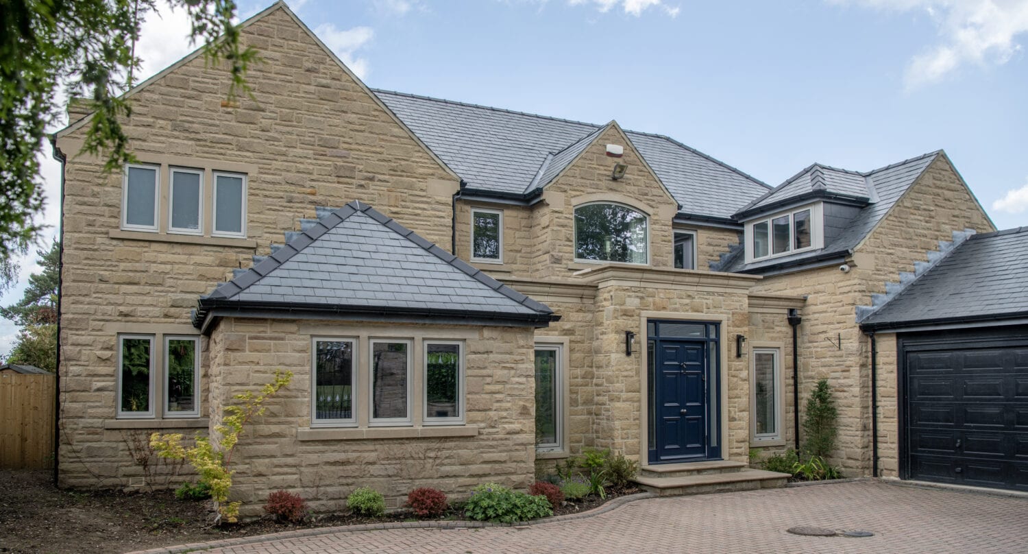 A modern two-story house with light stone walls, dark grey slate roof, large black front door, bespoke glass solutions in multiple windows, and a paved driveway surrounded by small shrubs and trees under a blue sky.
