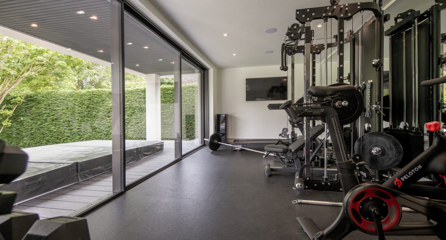 A modern home gym with various exercise equipment, including a weight machine, a stationary bike, and free weights, features large floor-to-ceiling windows and glass roofing overlooking a green outdoor area with a hot tub.