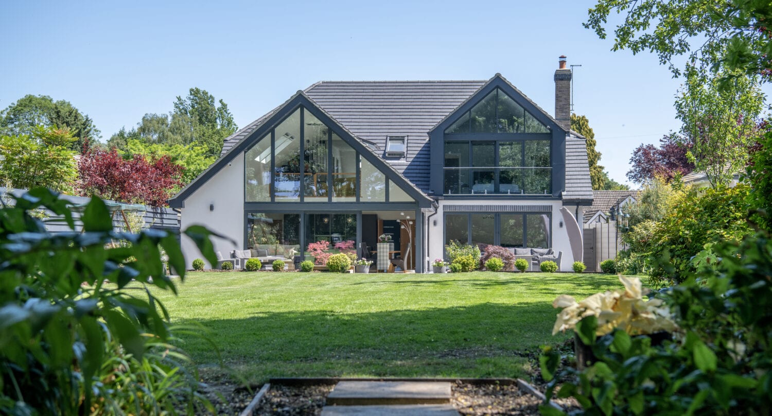 A modern two-story house with large glass windows and a sloped roof features sleek sliding doors, sitting in the center of a lush, green garden with a well-kept lawn, surrounded by trees and shrubs under a clear blue sky.