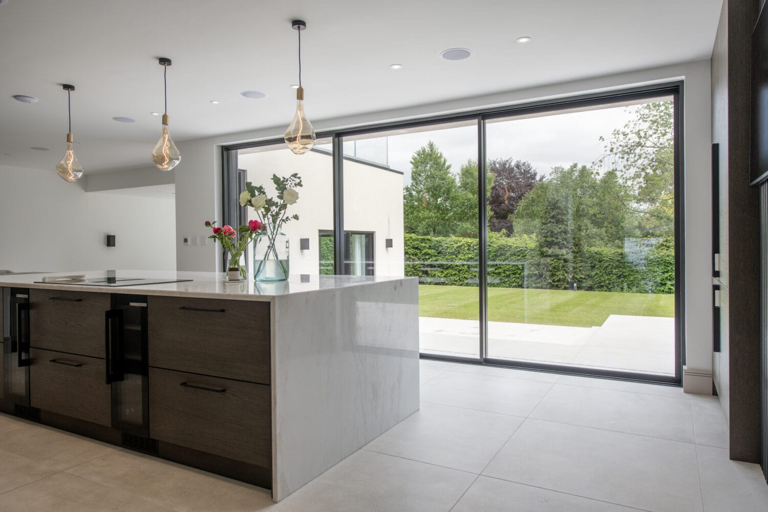 Modern kitchen with a marble island, pendant lights, and vases with flowers; large bifold doors open to a spacious green backyard with trees and lawn.