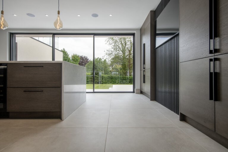 Modern kitchen with large gray floor tiles, wood cabinetry, an island, pendant lights, and sliding doors opening to a green garden with trees and hedges.