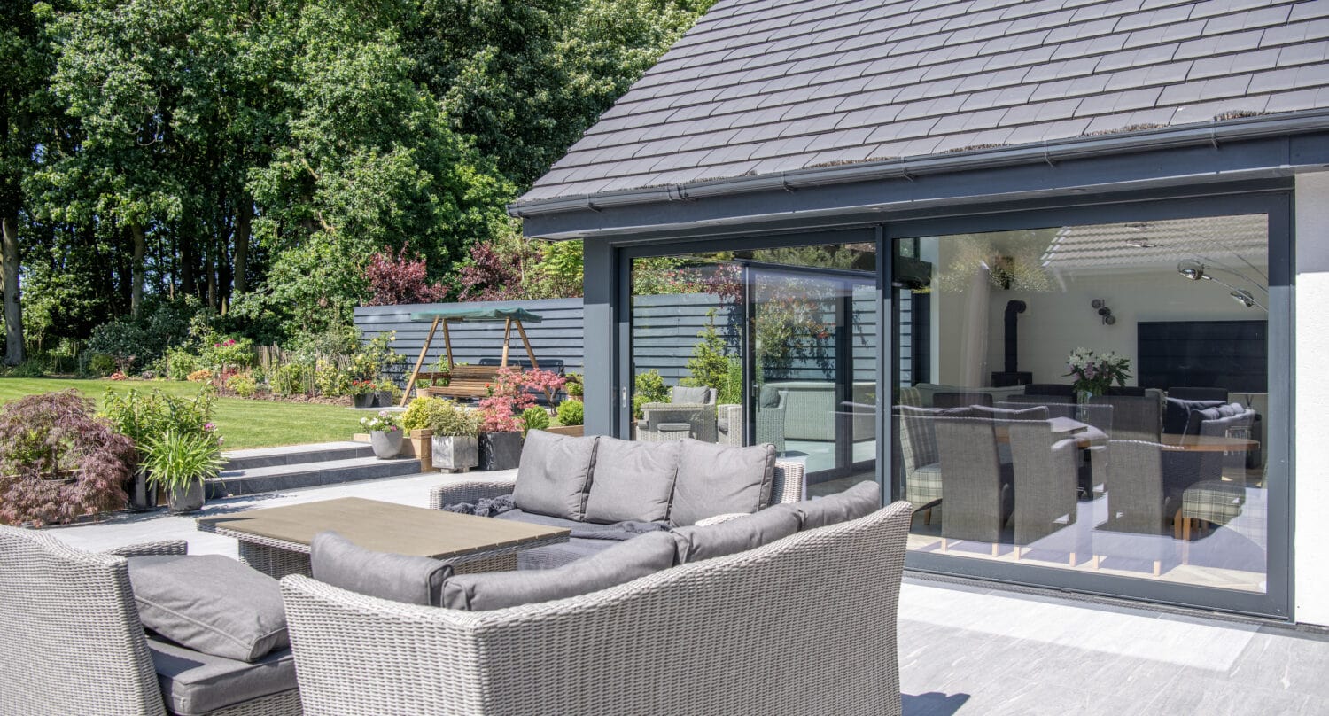 Modern patio with gray wicker sofas and table, set on a tiled terrace beside a house with large sliding doors. The garden features lush greenery, flowers, and a wooden swing beneath trees in the background. Consider adding glass roofing for year-round enjoyment.