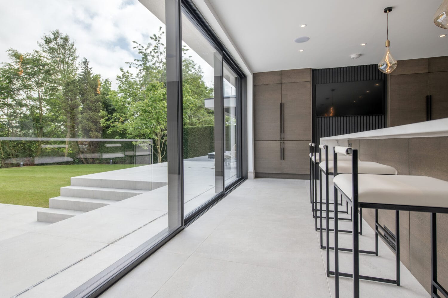 Modern kitchen with bar stools and large sliding doors opening to a patio and green lawn, featuring sleek cabinetry, minimalist design, and bespoke glass solutions, with outdoor steps leading to the garden.