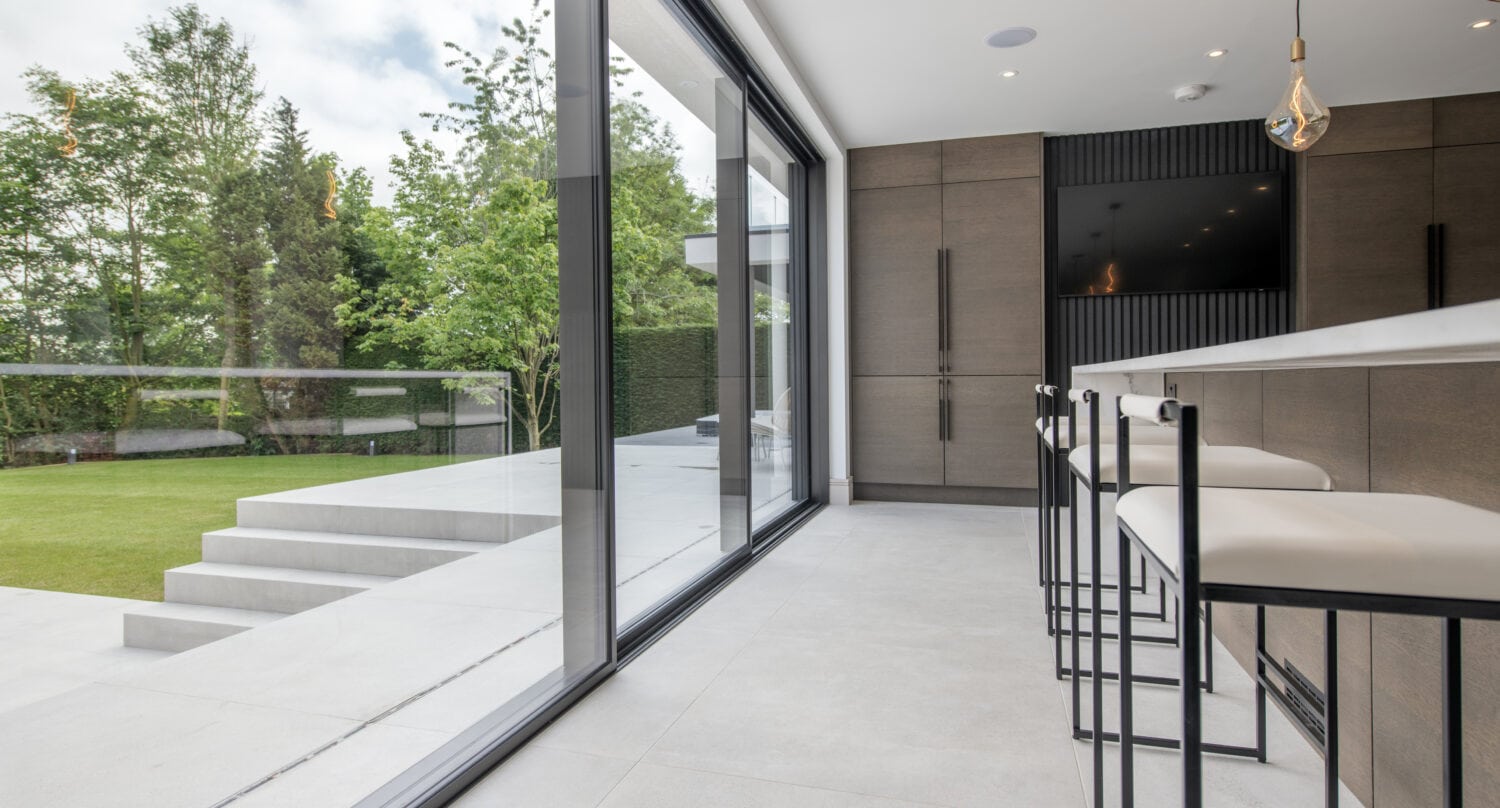 Modern kitchen with bar stools and large sliding doors opening to a patio and green lawn, featuring sleek cabinetry, minimalist design, and bespoke glass solutions, with outdoor steps leading to the garden.