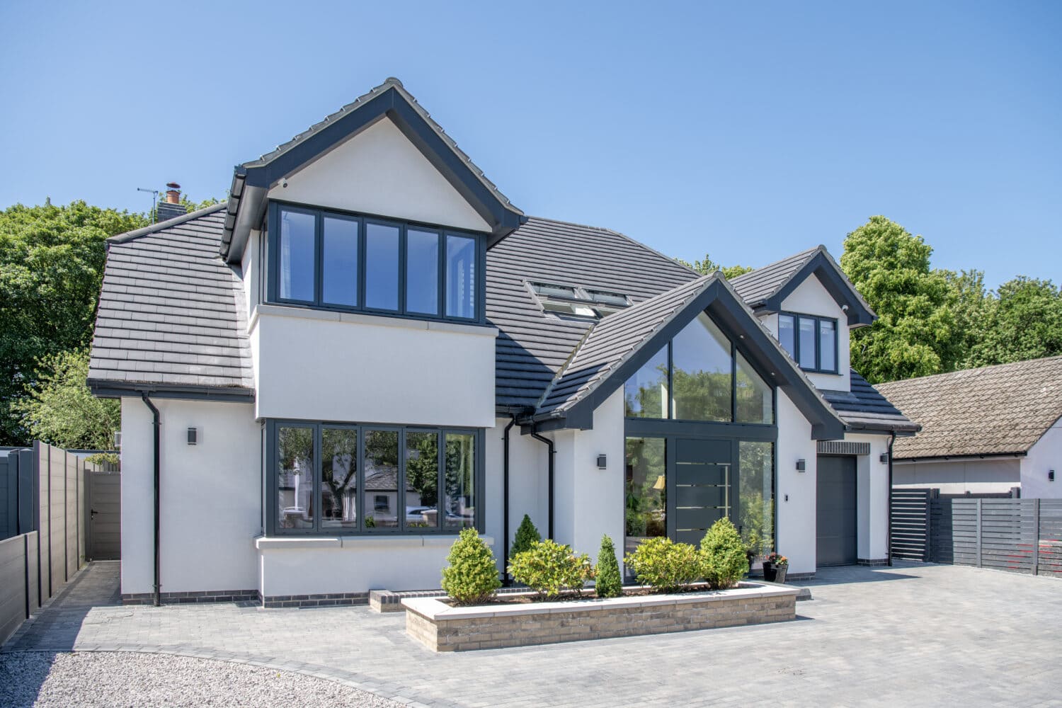 Modern two-story house with large windows, bespoke glass solutions, a gray roof, and white walls. Landscaped bushes line the front, with a paved driveway and trees in the background on a clear, sunny day.