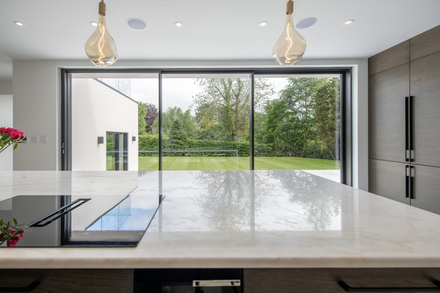 A modern kitchen with a marble island and built-in stovetop, hanging pendant lights, and large bifold doors overlooking a green garden and trees outside.