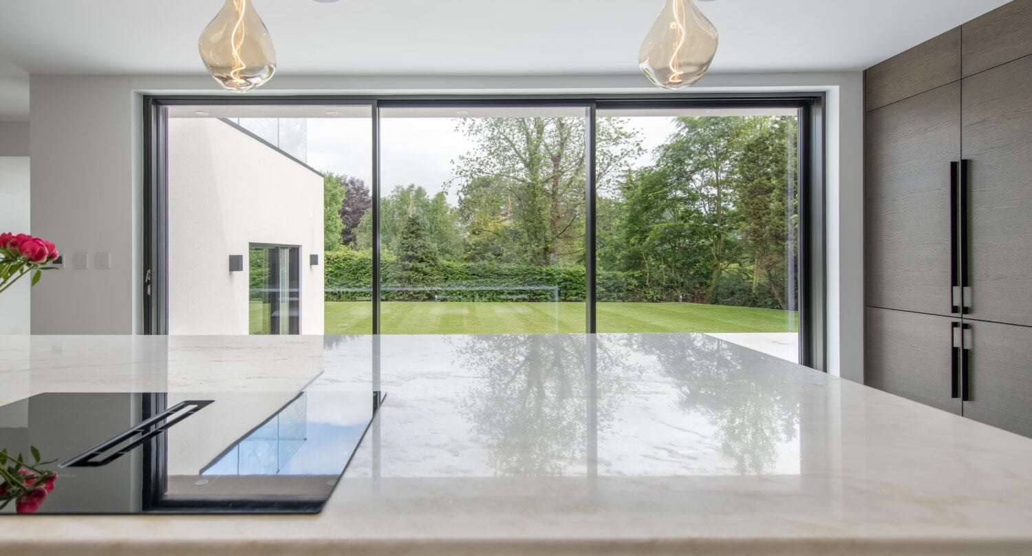 A modern kitchen with a marble island and built-in stovetop, hanging pendant lights, and large bifold doors overlooking a green garden and trees outside.