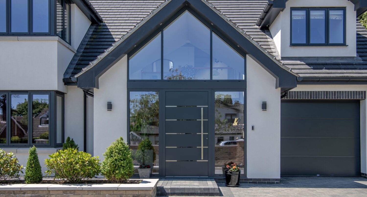 Modern house exterior with bespoke glass solutions, featuring a large glass front door, tall peaked roof, grey trim, symmetrical windows, a driveway, small garden beds with shrubs, and a single-car garage on the right.