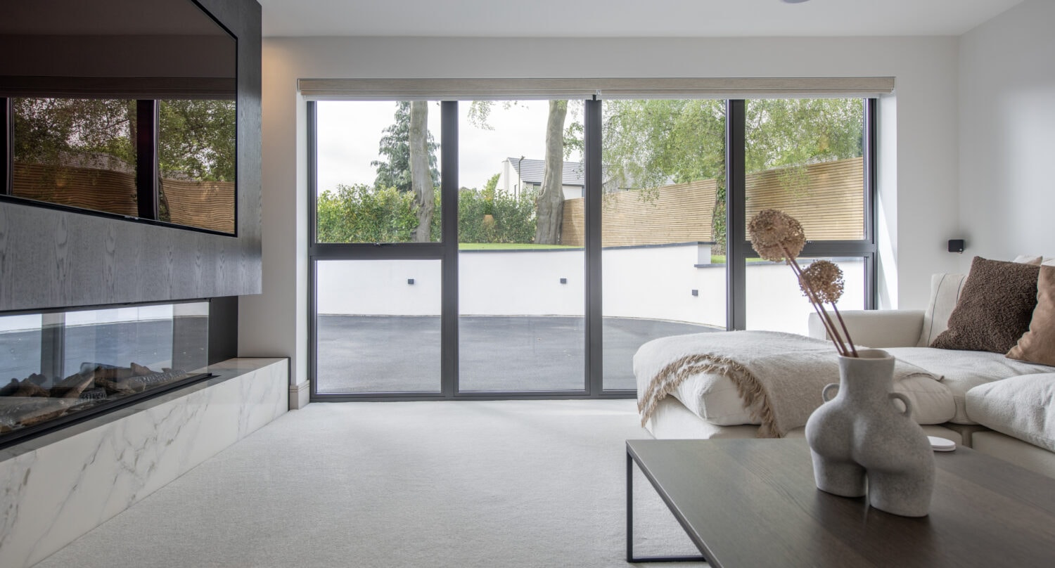 Modern living room with a large sectional sofa, sleek black and white fireplace, contemporary coffee table, and bespoke glass solutions in the floor-to-ceiling sliding doors overlooking a patio and lush greenery outside.