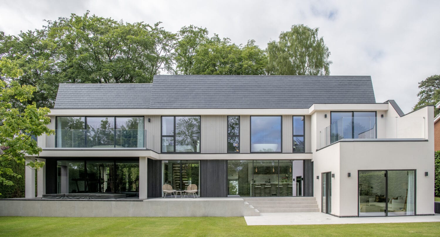 Modern two-story house with large windows, bespoke glass solutions, flat white exterior walls, and a dark sloped roof, surrounded by green grass and trees under a partly cloudy sky.