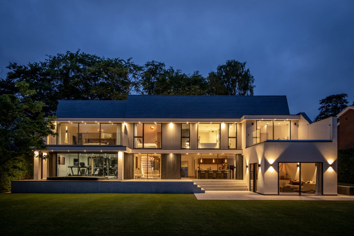 Modern two-story house at dusk with large glass windows, bespoke glass solutions like bifold doors and glass roofing, illuminated interior and exterior lights, a visible home gym, open living spaces, and a well-manicured lawn in the foreground.