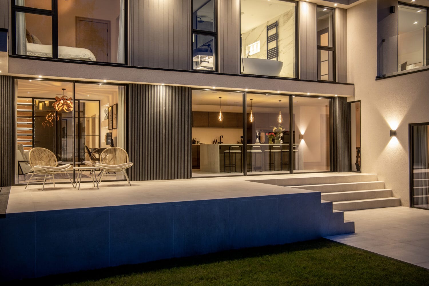 A modern house at dusk with large windows, sliding doors opening to a spacious patio, outdoor seating, and wide steps leading to a grassy yard. Warm lighting highlights the contemporary design.