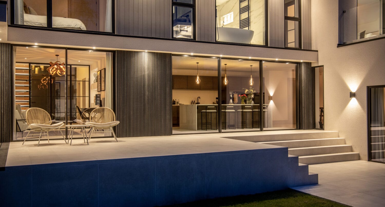 A modern house at dusk with large windows, sliding doors opening to a spacious patio, outdoor seating, and wide steps leading to a grassy yard. Warm lighting highlights the contemporary design.