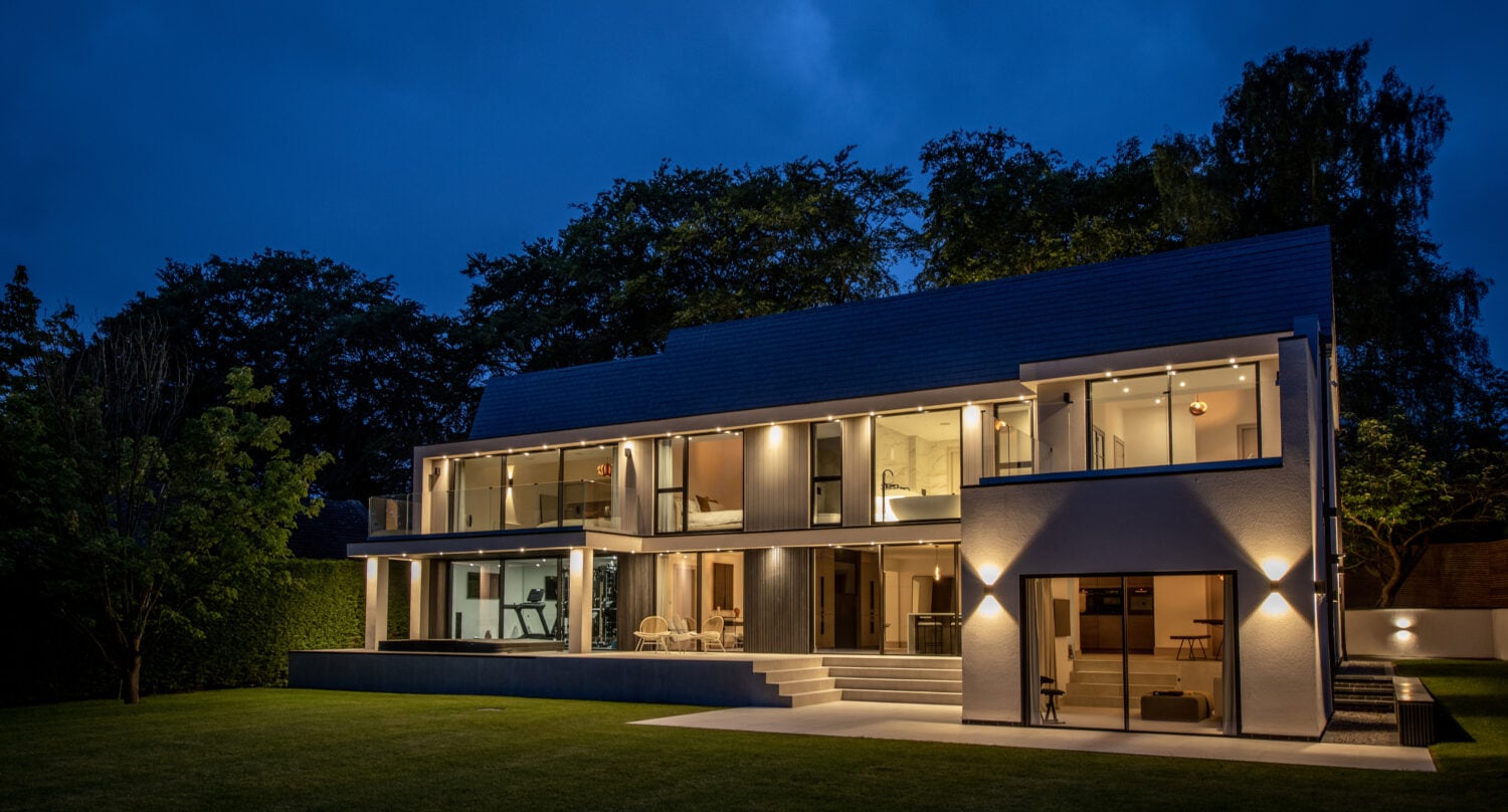 A modern two-story house with large windows and bright interior lights, viewed at dusk. Sliding doors open to a well-kept lawn, while bespoke glass solutions enhance the homes sleek, contemporary design amid surrounding trees.