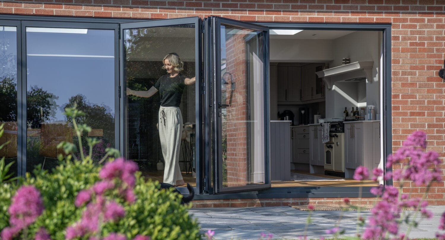 A person stands smiling in the doorway of a modern brick house with large bifold doors open to a garden filled with green bushes and bright pink flowers. The kitchen is visible inside.