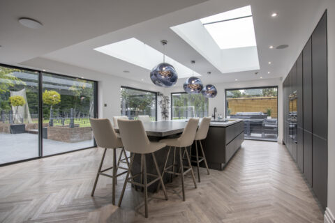 Modern kitchen with large island, four barstools, and three pendant lights. Skylights and floor-to-ceiling sliding doors fill the space with natural light. Outdoor patio and garden are visible through the expansive sliding glass doors.
