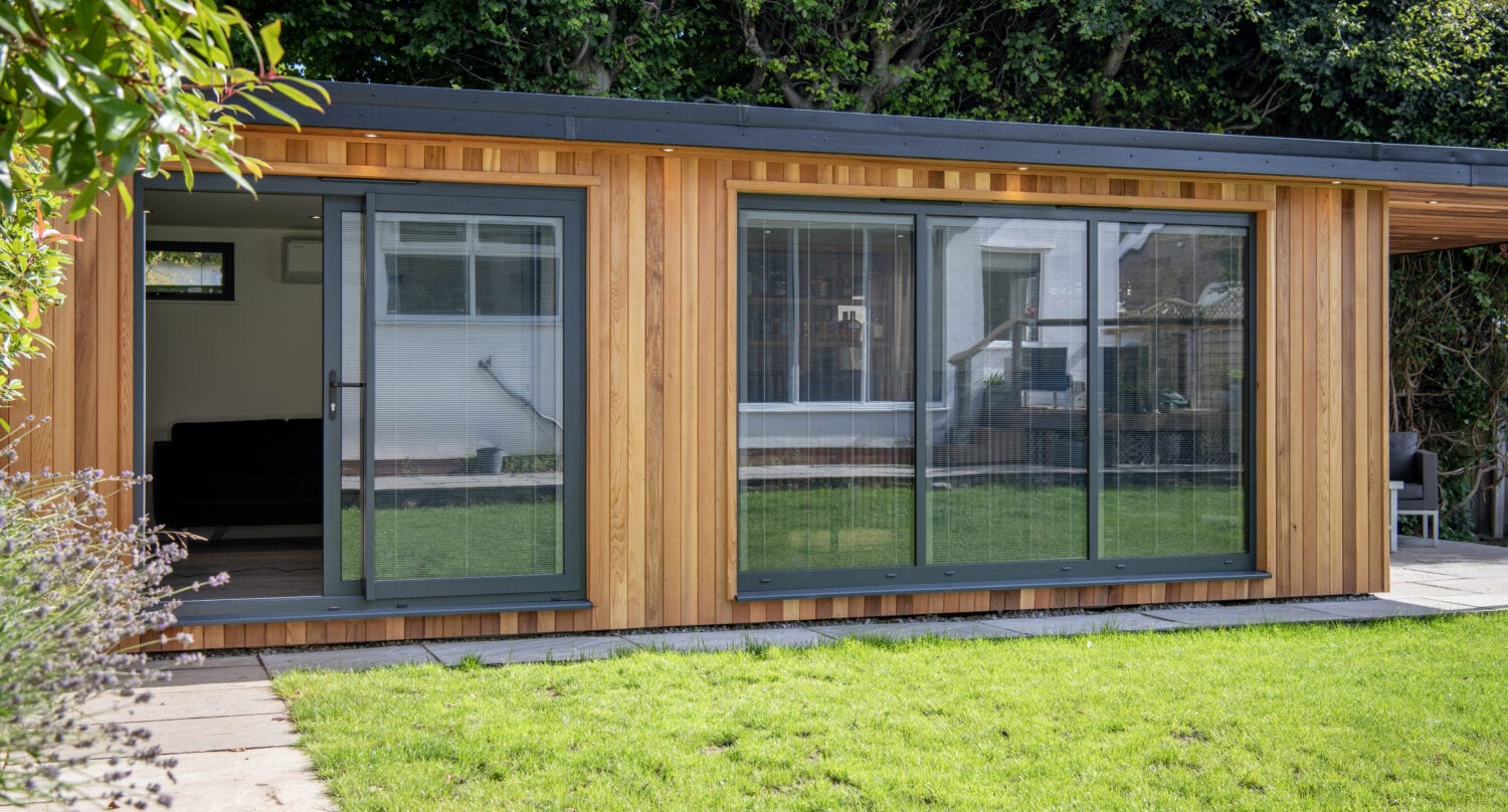 A modern wooden garden room with large sliding glass doors, set on a patio and surrounded by green grass and hedges. Reflections show a nearby house, highlighting bespoke glass solutions for seamless indoor-outdoor living.