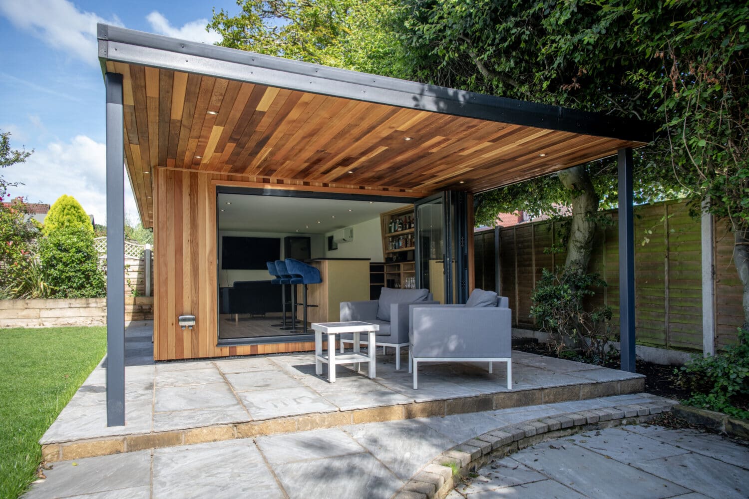 Modern garden room with a wooden ceiling and open bifold doors, featuring outdoor seating on a stone patio, surrounded by greenery and a wooden fence—a showcase of bespoke glass solutions.