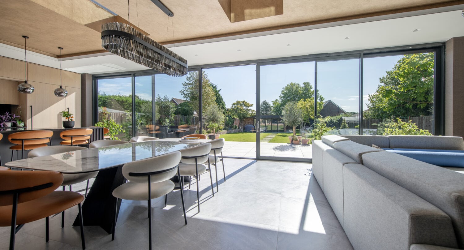 Modern dining and living area with bespoke glass solutions, including large sliding doors opening to a sunny garden. The room features a long dining table with chairs, a contemporary chandelier, kitchen island, and light gray sectional sofa.