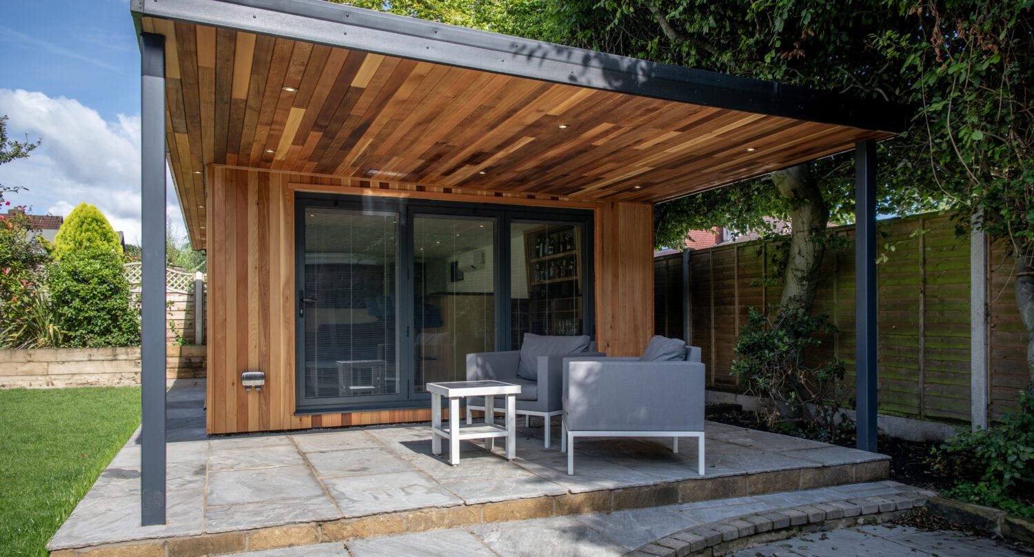 Modern wooden garden room with bifold doors, a covered patio area, and gray outdoor furniture, set on a paved area surrounded by grass, trees, and a wooden fence.
