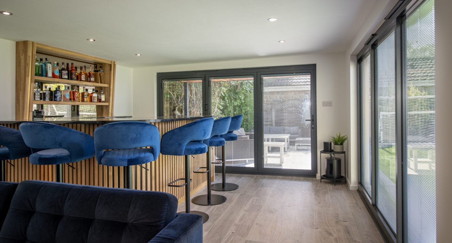A modern home bar with blue velvet bar stools, a wooden bar counter, and shelves of liquor bottles. Large sliding doors let in natural light, and there’s a small potted plant by the entrance to a patio area.
