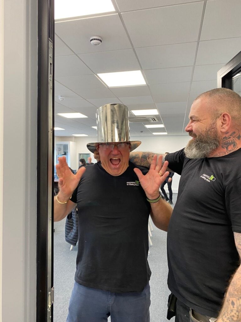 A man wearing a shiny, metallic top hat stands in a doorway making a playful face with hands up, while another man with tattoos on his arm stands beside him, smiling. Both are in a modern office with stylish bifold doors.