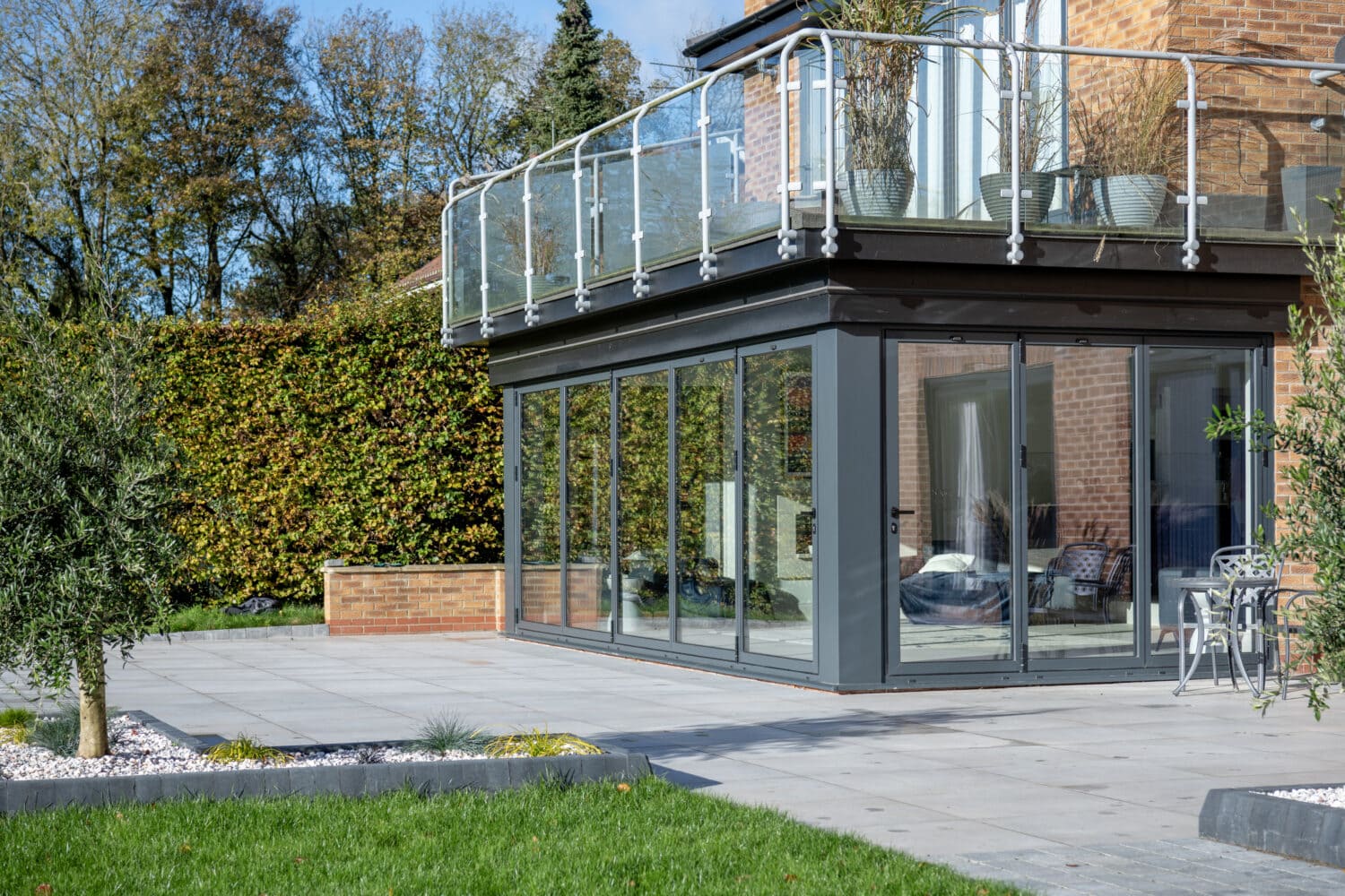 A modern house extension with large bifold doors and a glass balcony above, surrounded by a paved patio, greenery, and a well-kept lawn.