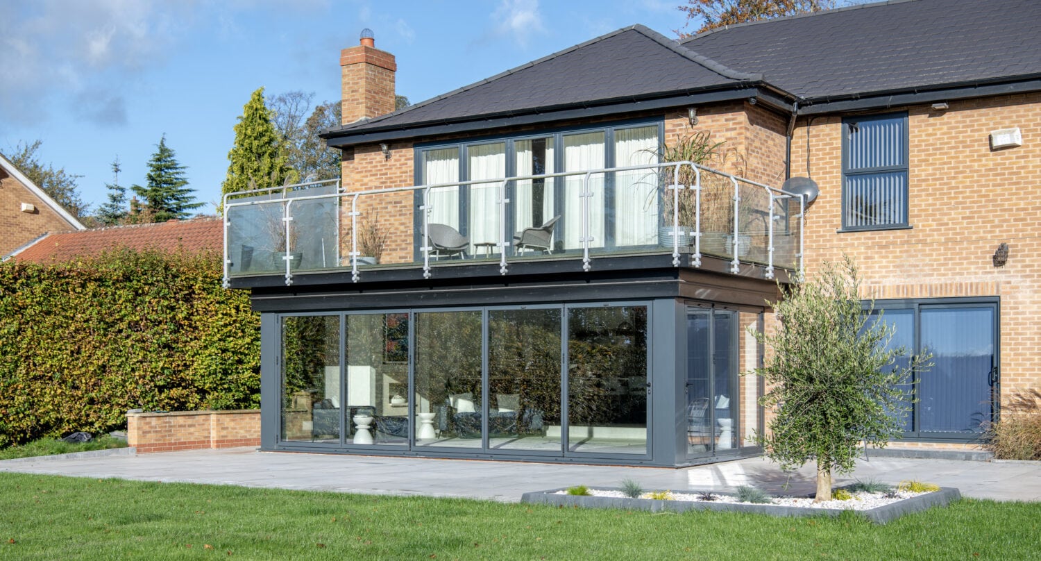A modern two-story brick house with large floor-to-ceiling windows, sleek sliding doors, a glass balcony, and a neatly landscaped yard featuring a small tree and green lawn under a blue sky.