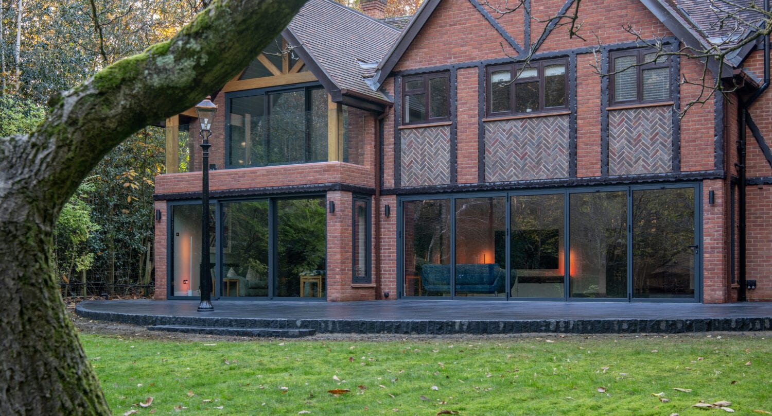 A modern red-brick house with large bespoke glass solutions, including bifold doors and expansive windows. Warm lights glow inside, and the home sits among green grass, trees, and a mossy tree trunk in the foreground.