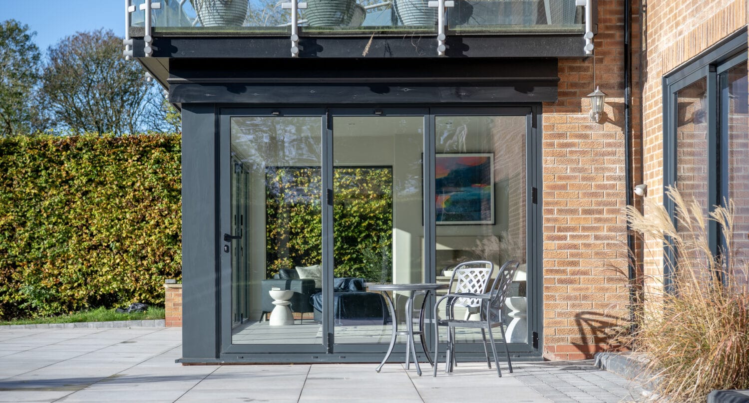 Modern glass-enclosed sunroom with bespoke glass solutions attached to a brick house, featuring a small patio table and two chairs on a paved terrace, plus large potted plants on the balcony above.