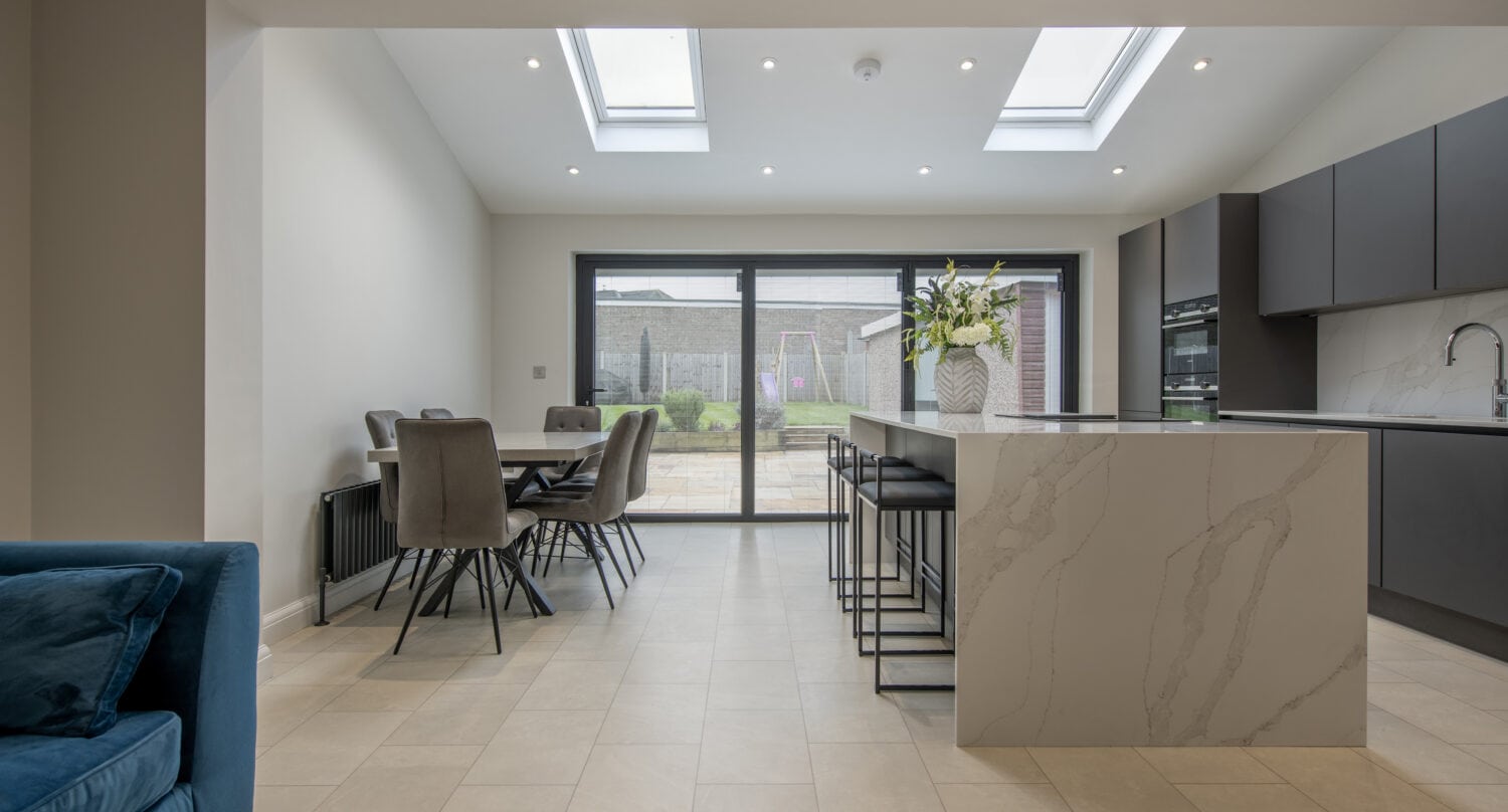 Modern kitchen and dining area with marble island, gray cabinets, and skylights. Large sliding doors open to a patio and garden. Neutral tones and elegant, contemporary furnishings complete the space with bespoke glass solutions for a refined touch.