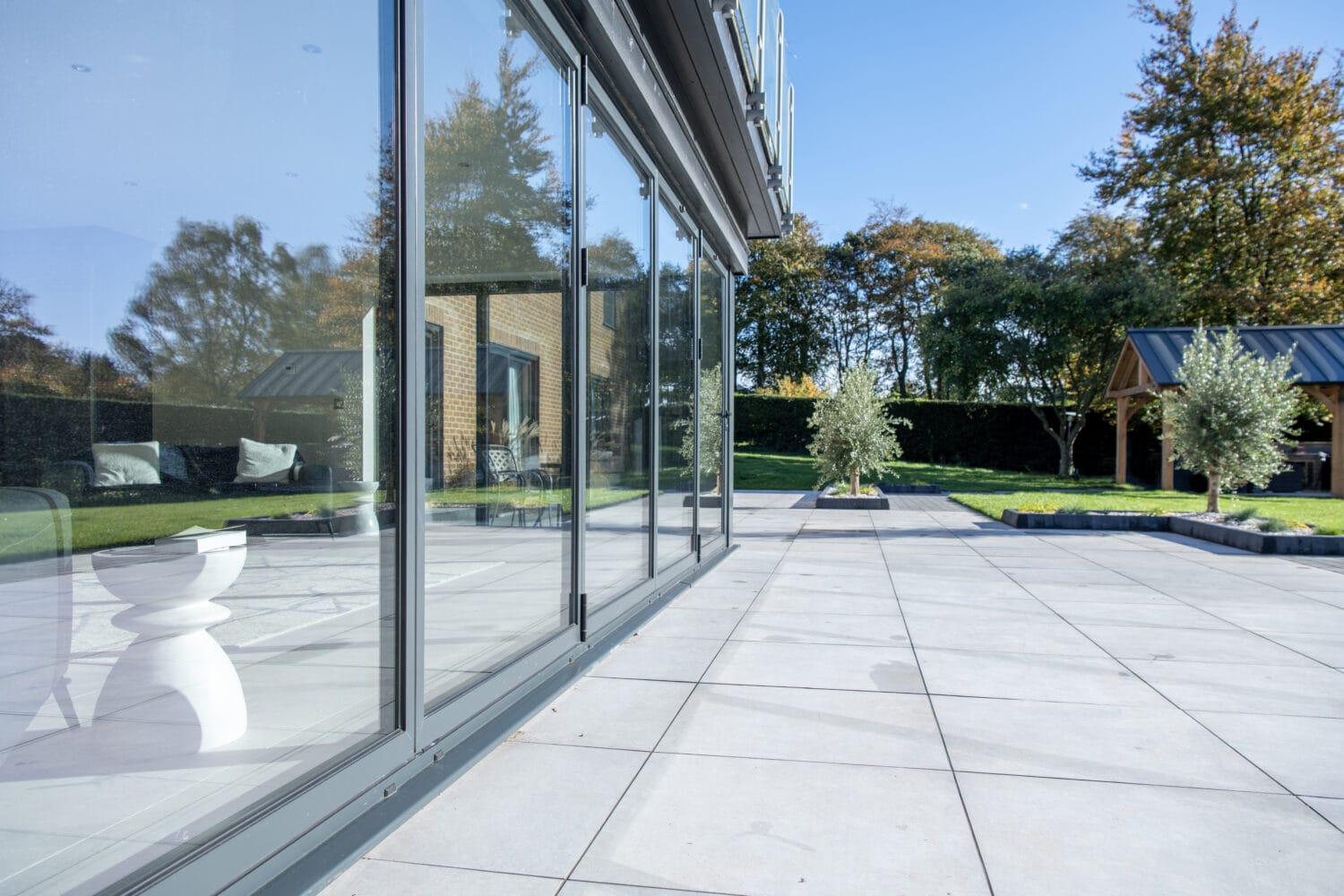 A modern patio with large glass sliding doors and bespoke glass solutions reflects trees and sky. The area is paved with light gray tiles and opens onto a green lawn with trees and a wooden gazebo in the background.