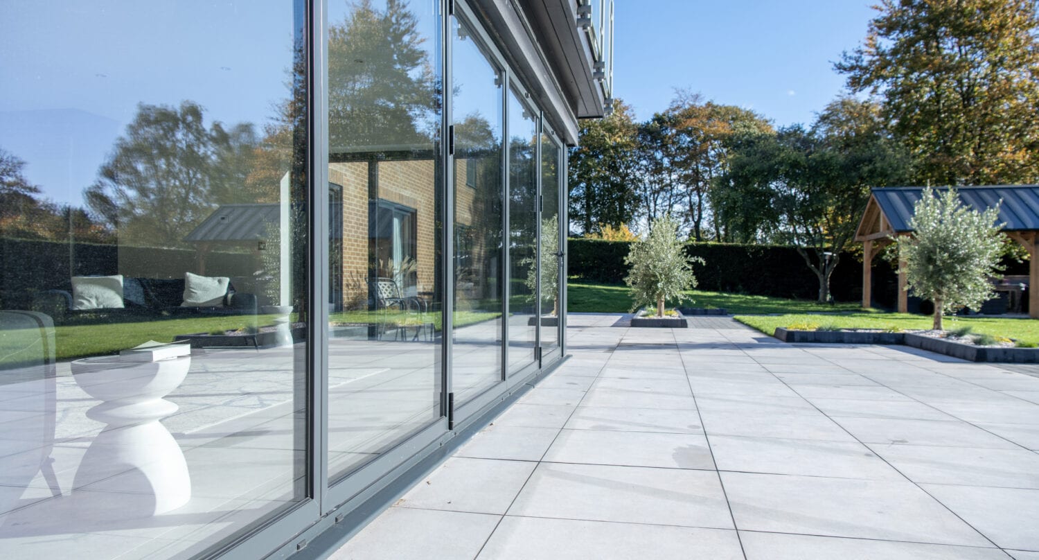 A modern patio with large glass sliding doors and bespoke glass solutions reflects trees and sky. The area is paved with light gray tiles and opens onto a green lawn with trees and a wooden gazebo in the background.
