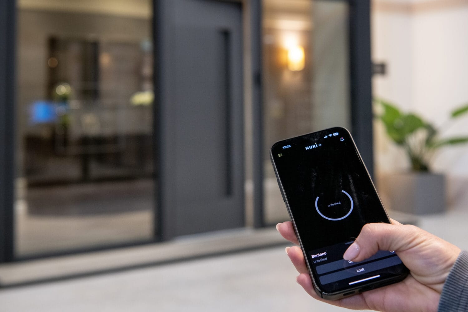 A person holds a smartphone with an app open, appearing to unlock sliding doors remotely. The background shows a modern entrance with glass doors and indoor plants.