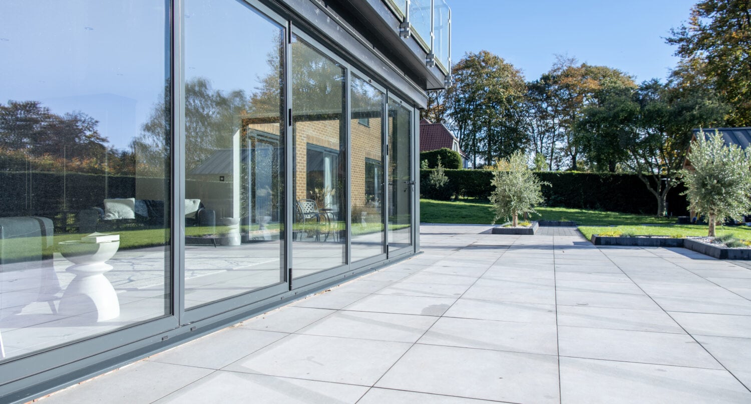 Large glass sliding doors open onto a spacious modern patio with light gray tiles and lush lawn, while glass roofing overhead lets in natural light, surrounded by trees and a clear blue sky in the background.