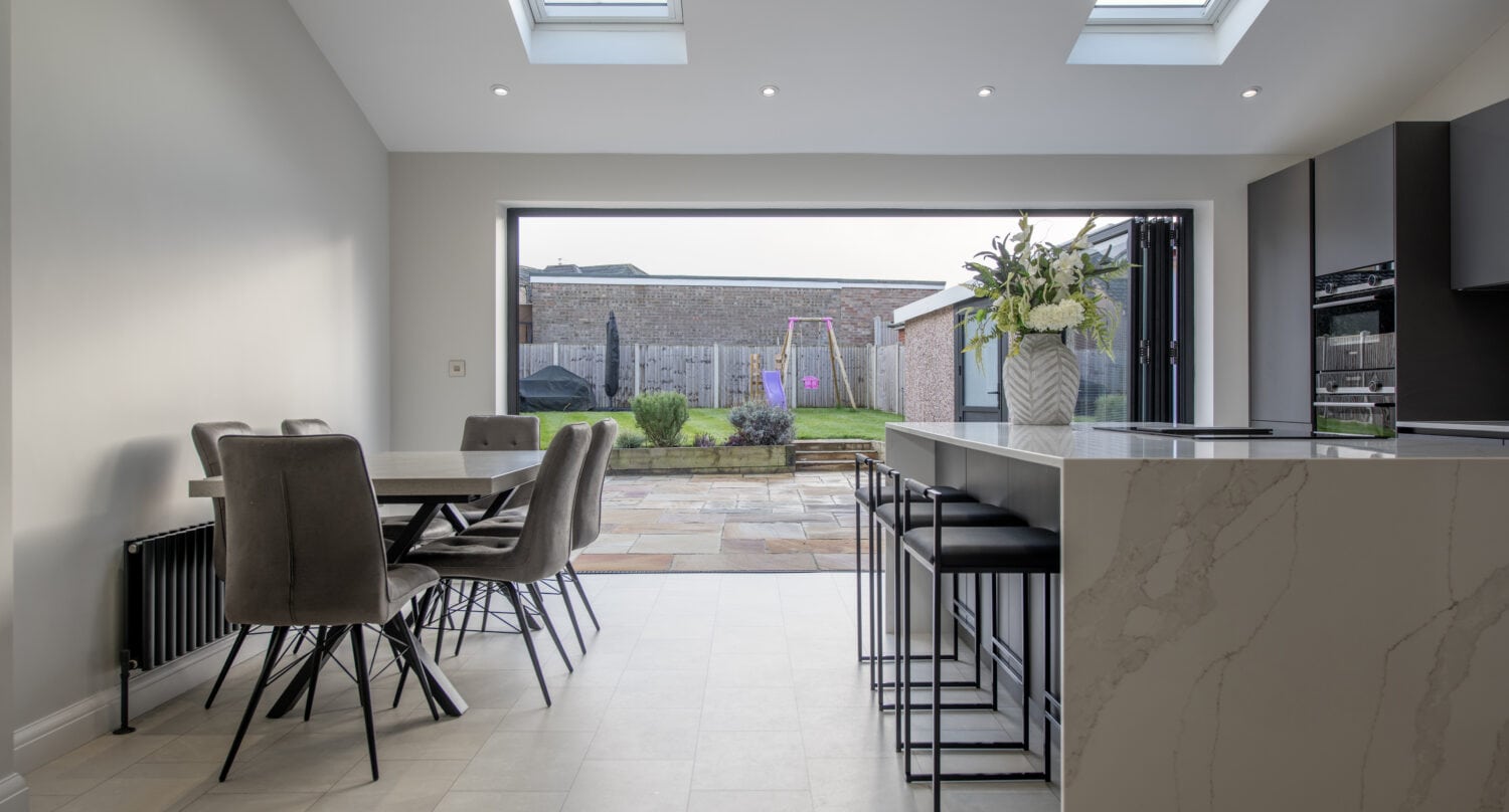 Modern kitchen and dining area with marble island, gray chairs, and large skylights. Glass roofing and sliding doors open to a patio and fenced backyard with a swing set and garden. Natural light fills the space.