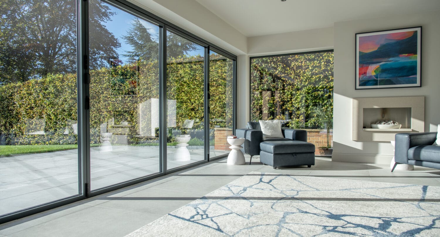 A modern living room with large bifold doors opening to a patio, two dark armchairs, a patterned rug, abstract wall art, and greenery visible outside. Sunlight streams into the bright space through bespoke glass solutions.