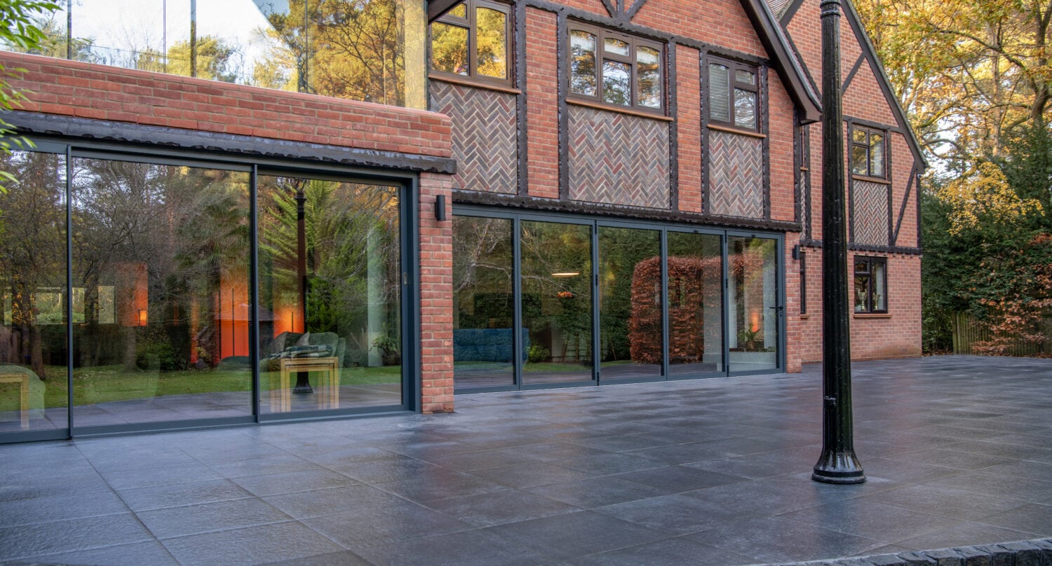 A modern brick house with large glass sliding doors and windows, reflecting trees and greenery. The spacious patio features dark tiles, a vintage-style black lamp post on the right, and autumn leaves scattered around.