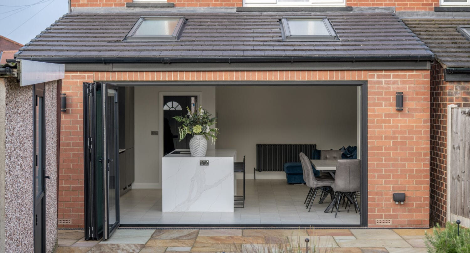 Modern brick house extension with large sliding doors reveals a bright kitchen and dining area. The space features a white island with a vase of flowers, grey upholstered chairs, and bespoke glass solutions that flood the interior with natural light.