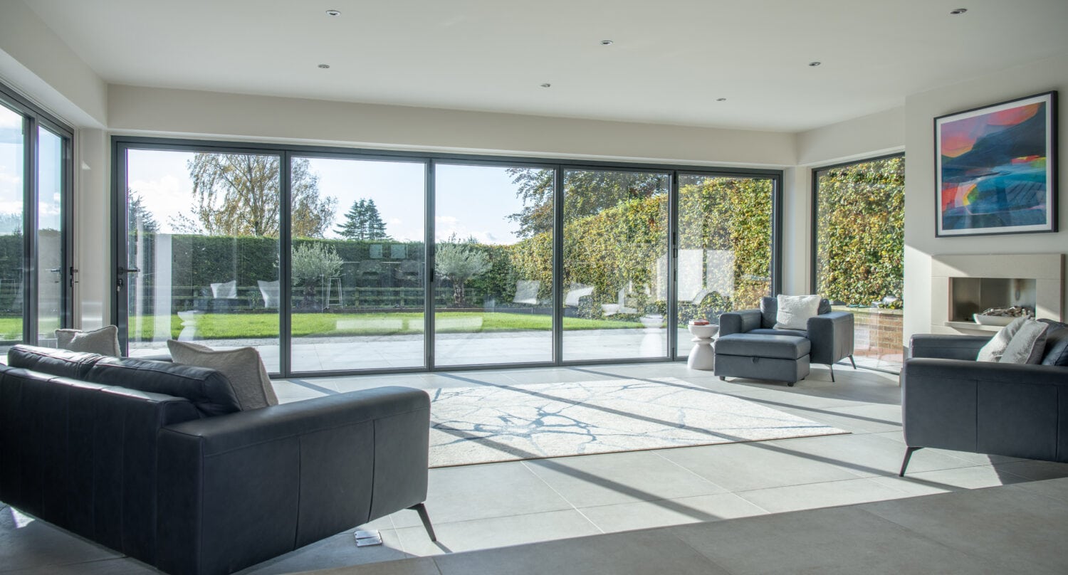 Modern living room with large floor-to-ceiling sliding doors overlooking a garden. The room features dark sofas, a rug, an armchair, and wall art above a fireplace. Sunlight from the bespoke glass solutions fills the bright, open space.