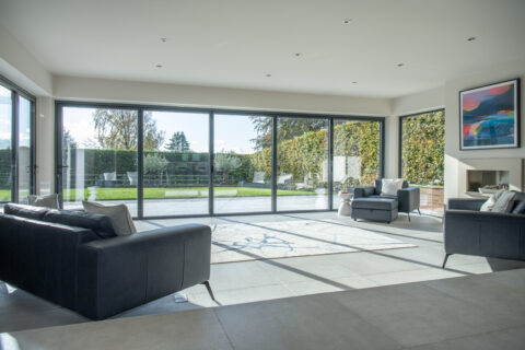 Modern living room with large floor-to-ceiling sliding doors overlooking a garden. The room features dark sofas, a rug, an armchair, and wall art above a fireplace. Sunlight from the bespoke glass solutions fills the bright, open space.