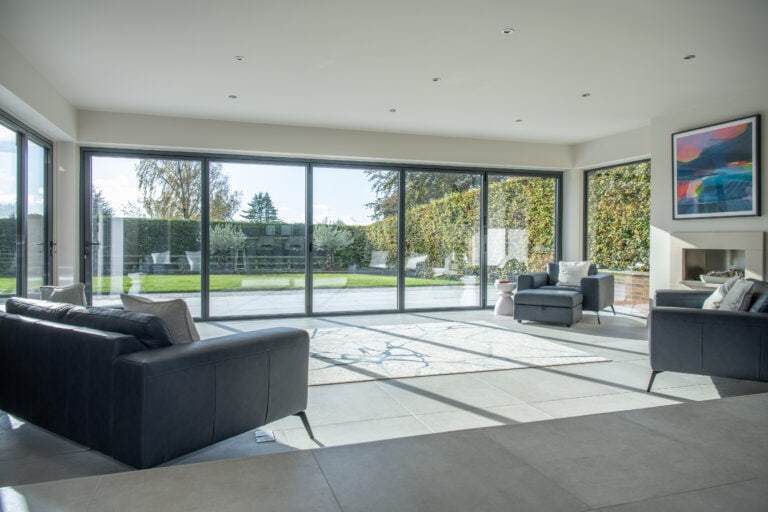 Modern living room with large floor-to-ceiling sliding doors overlooking a garden. The room features dark sofas, a rug, an armchair, and wall art above a fireplace. Sunlight from the bespoke glass solutions fills the bright, open space.