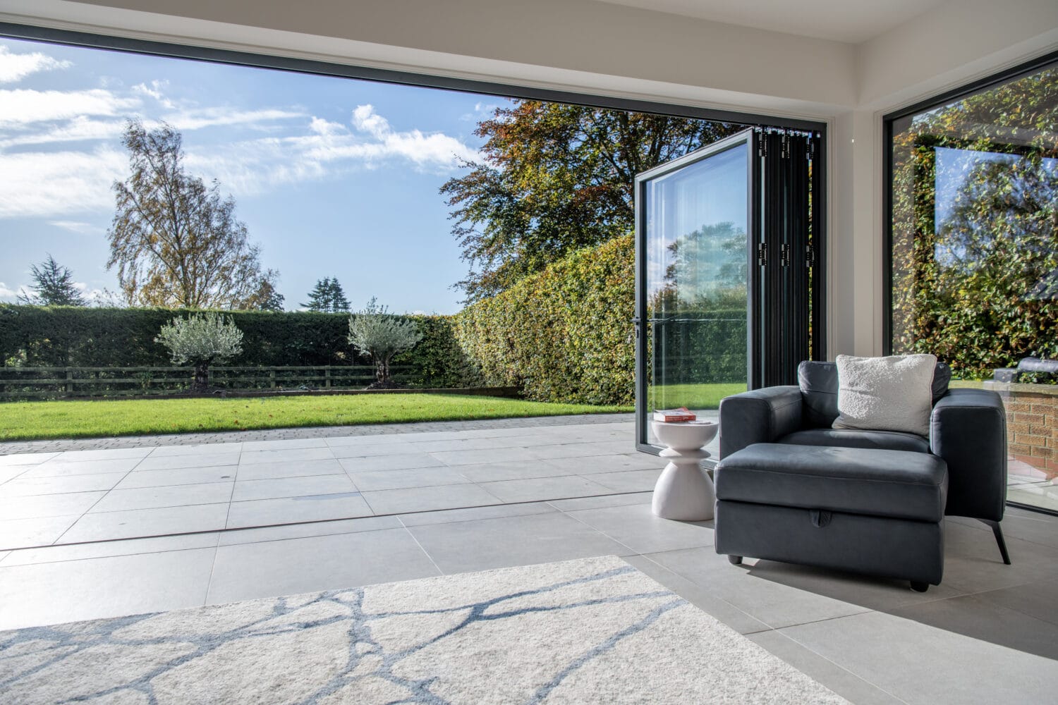 A modern living space with large bifold doors open to a green garden; a dark armchair and small white side table sit on tiled flooring near a gray patterned rug. Trees and a blue sky are visible outside.