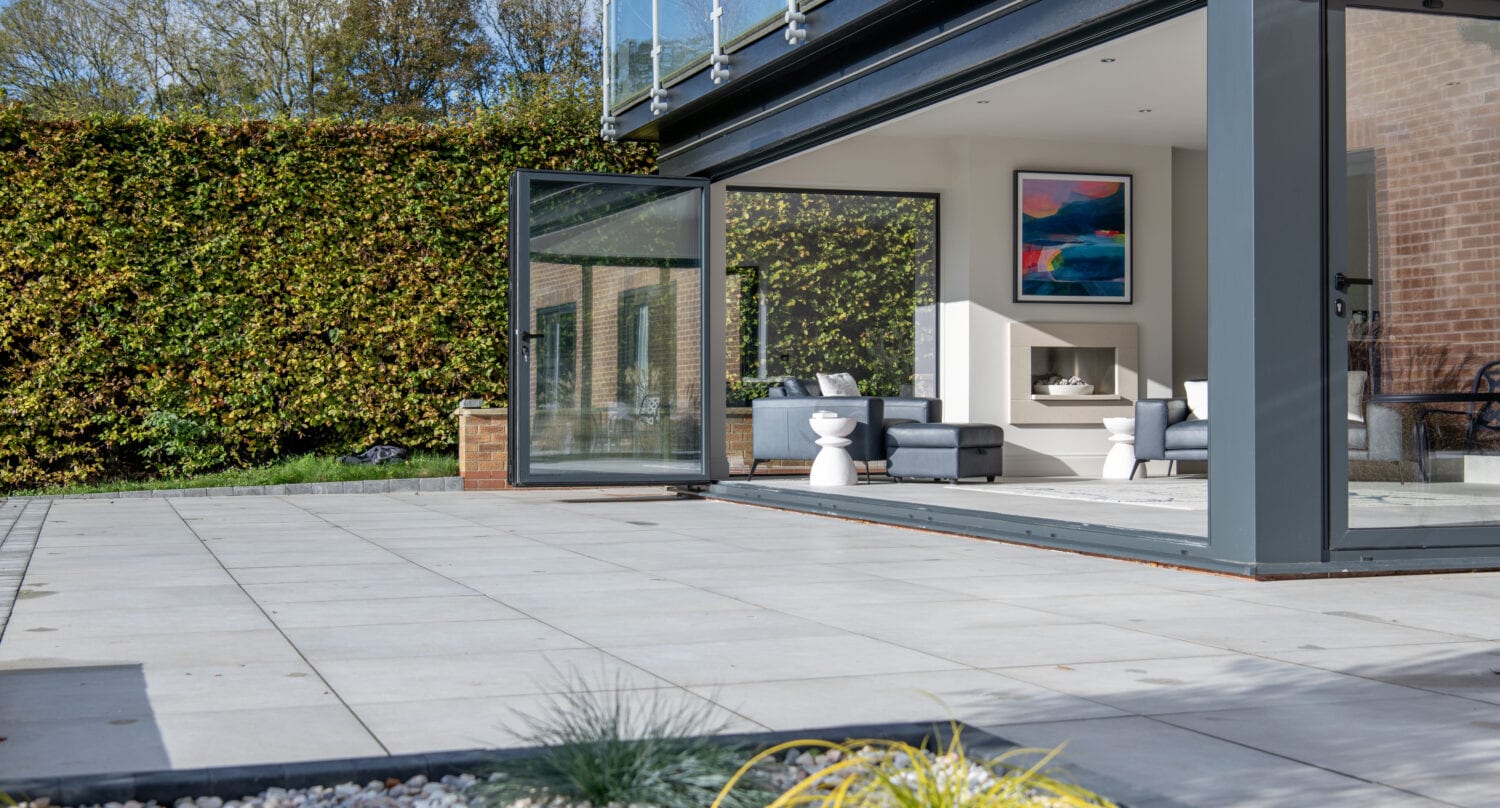 Modern house with large bifold doors open to a spacious patio. Inside, a light gray sofa, white stool, fireplace, and abstract wall art add style. The tiled patio features a rock and plant garden in the foreground.