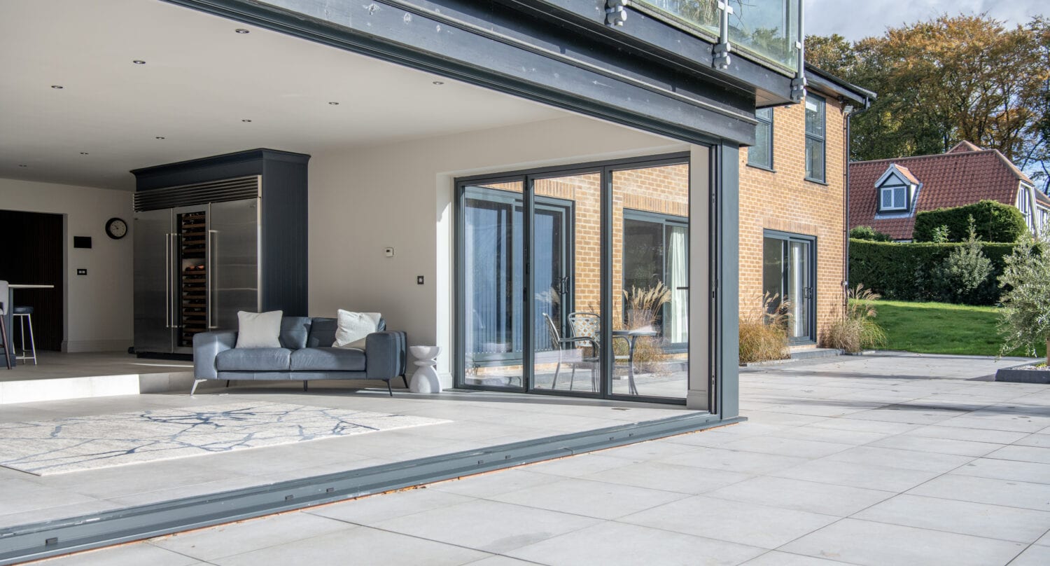 A modern house with large bifold doors opens to a spacious patio, revealing a living area with a gray sofa and rug, and an outdoor area with greenery and a neighboring house in the background.