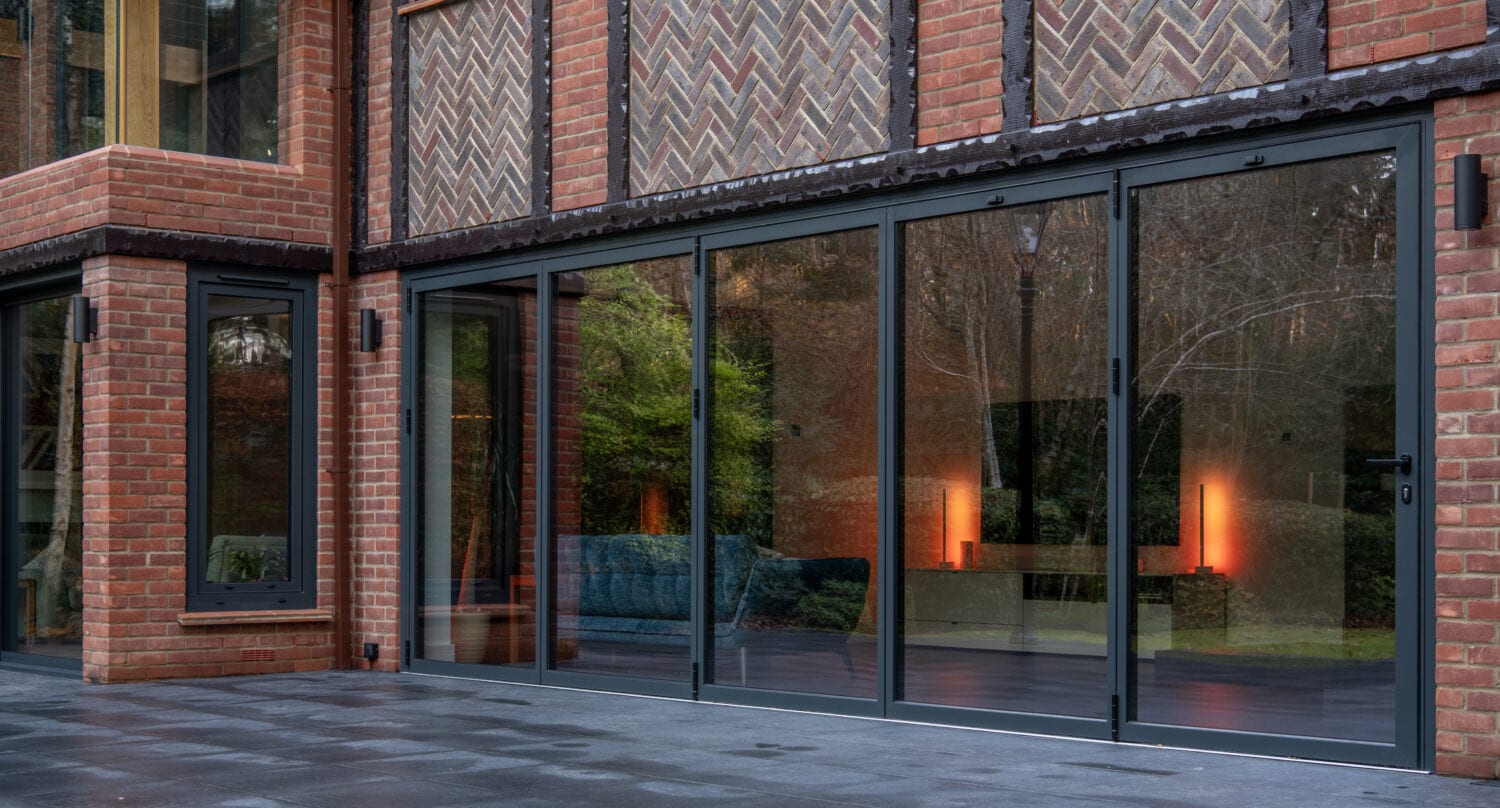 Large bifold doors on a modern brick house open to a tiled patio; warm interior lights and reflections of trees are visible through the bespoke glass solutions.