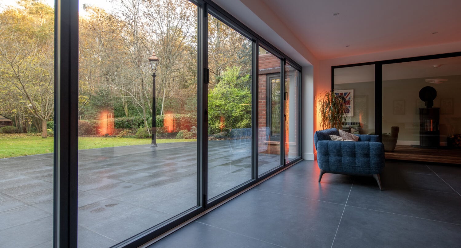 A modern living room with large bifold doors overlooking a spacious patio and green garden. Inside, a blue armchair and contemporary decor are complemented by natural light filling the room, thanks to bespoke glass solutions.